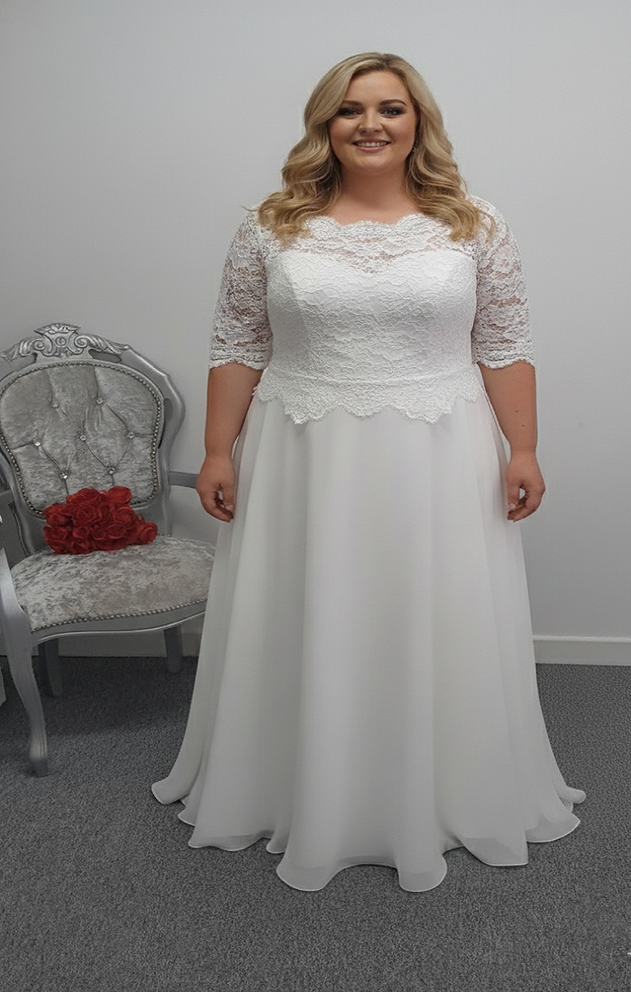 Woman wearing a white lace wedding dress in a room with a chair and red flowers.